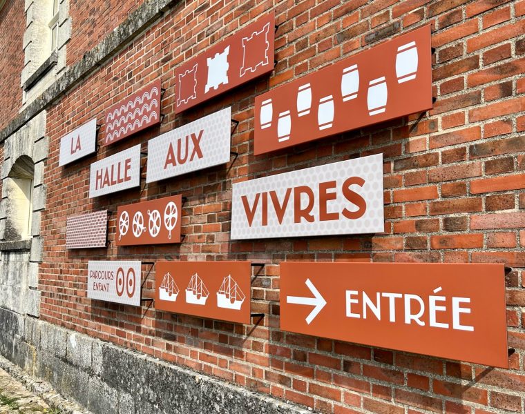 panneaux directionnels oranges fixés au mur de pierre de la Halle aux Vivres de Brouage. Les panneaux indiquent le chemin vers l'exposition permanente et d'autres points d'intérêt du site.